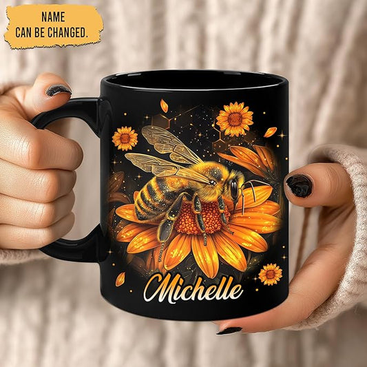 Black mug with bee and sunflower artwork and script name Michelle, held by hands