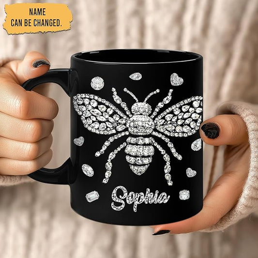 Black coffee mug held by hands; rhinestone-style bee design with name Saphia