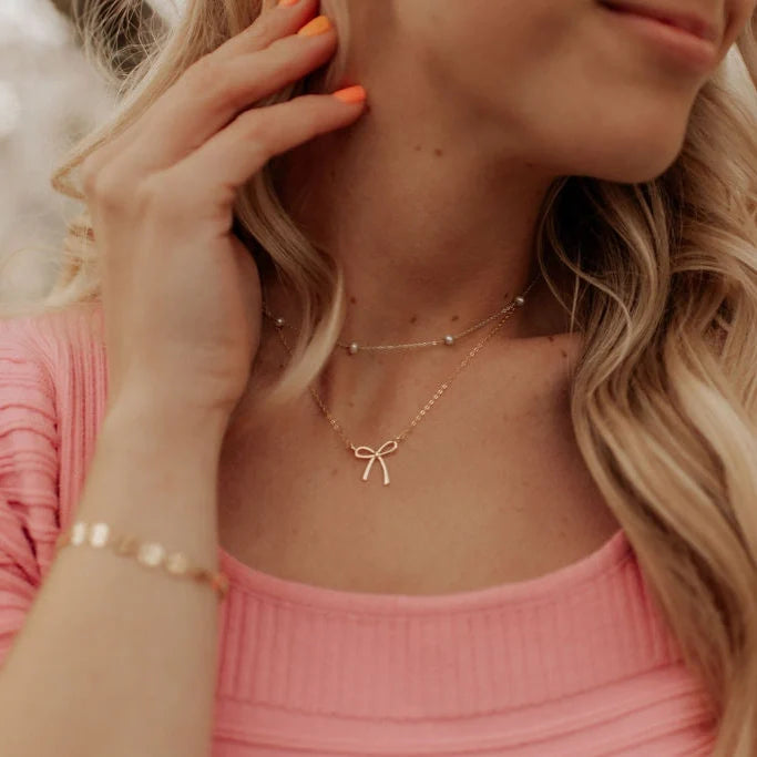 Gold bow pendant necklace shown layered with a beaded choker