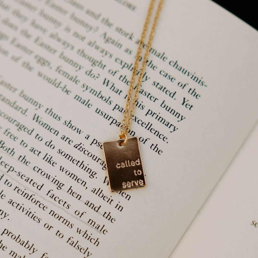 Gold rectangular Called to Serve necklace with dark reflective lettering.