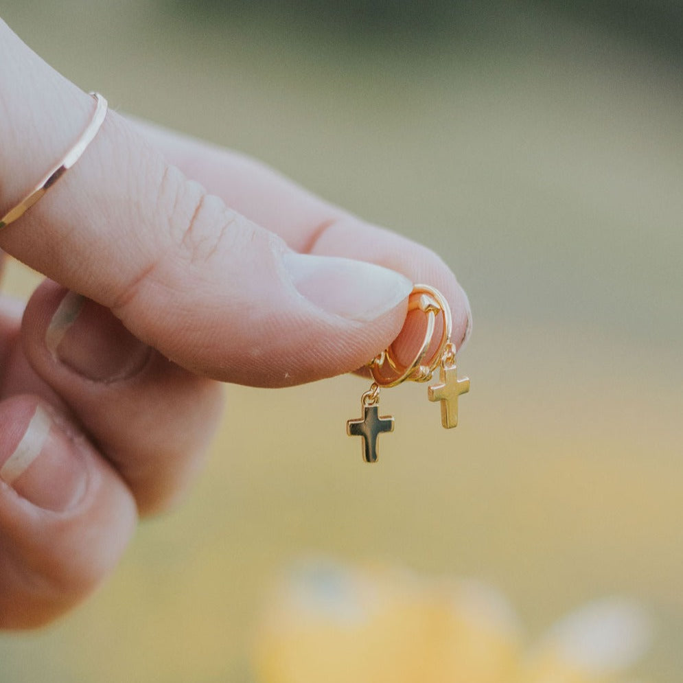 Gold-tone hoop earrings with dangling cross charms held between fingers