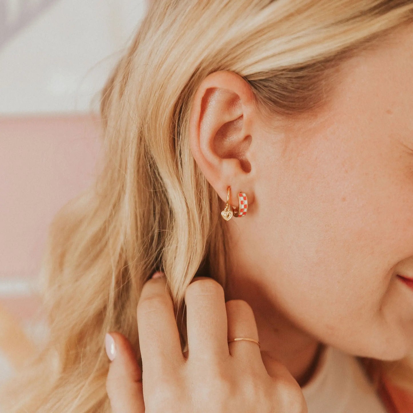 Small gold hoop with a dangling heart charm, worn beside a red checkerboard hoop