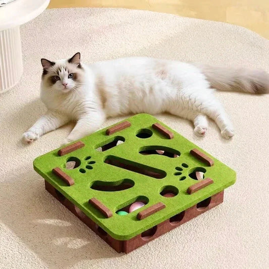 Cat lounging beside green felt maze puzzle box with paw-print cutouts and ball holes.