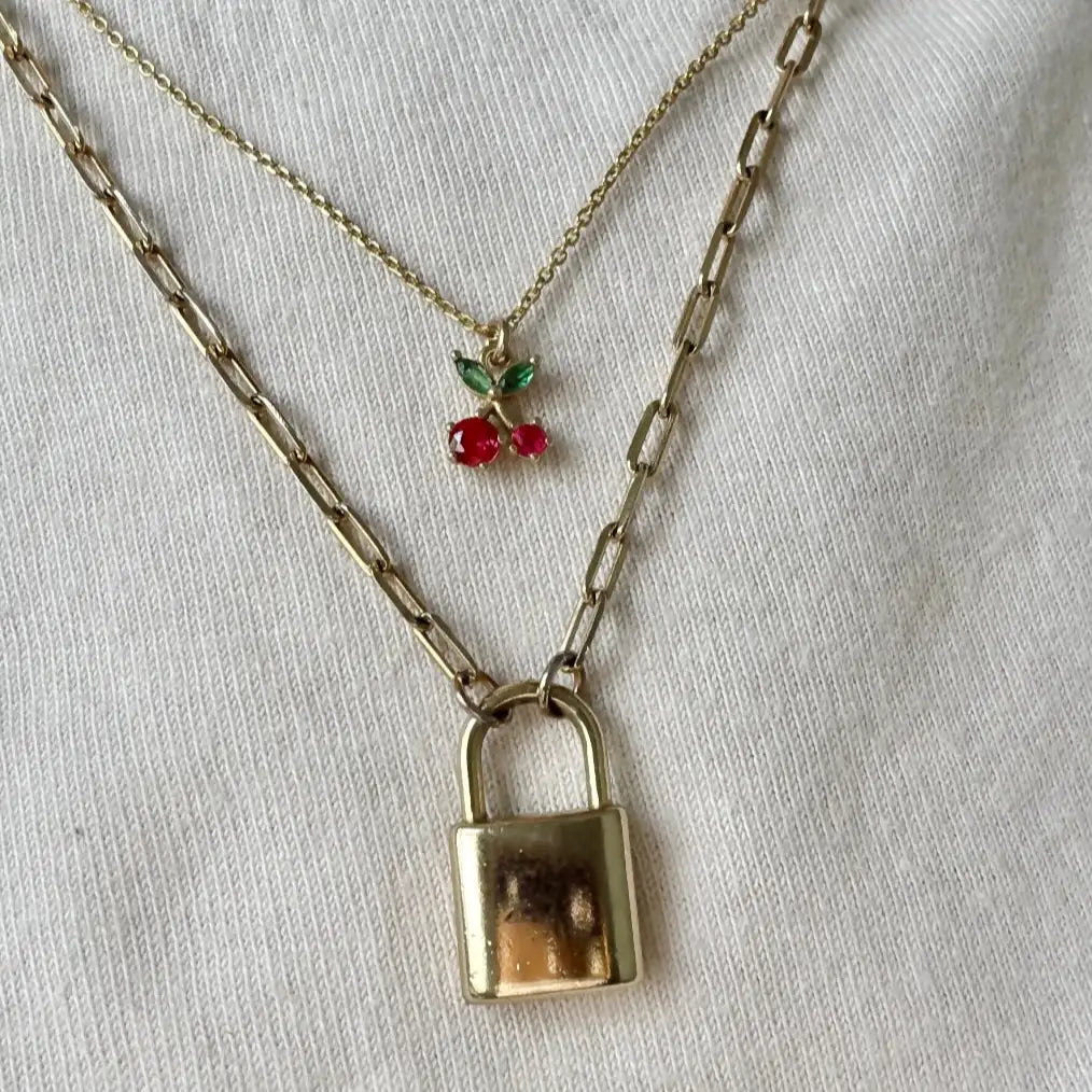 Delicate gold cherry necklace with red and green stones, layered above a padlock necklace