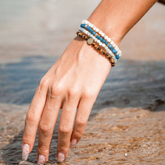 Four beaded bracelets on wrist: brown, white, and blue bead stacks with gold charm