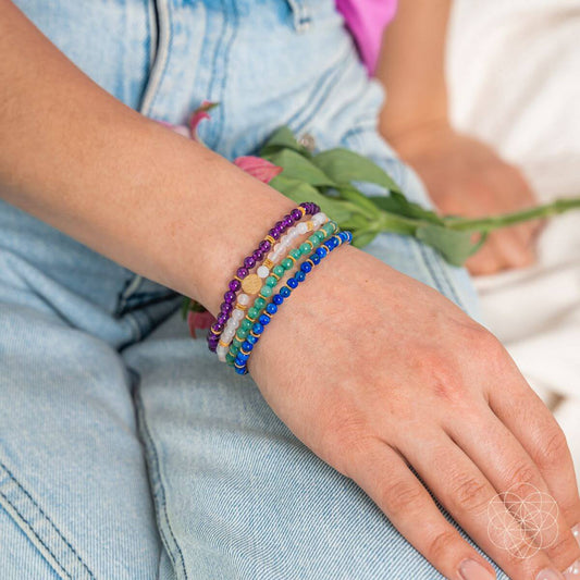 Four beaded charm bracelets worn on wrist, featuring purple, blue, green, and white beads