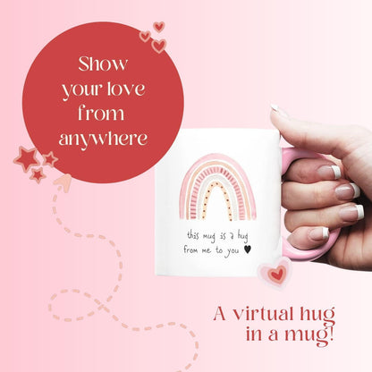 White ceramic mug with pink handle and rainbow hug quote card being held