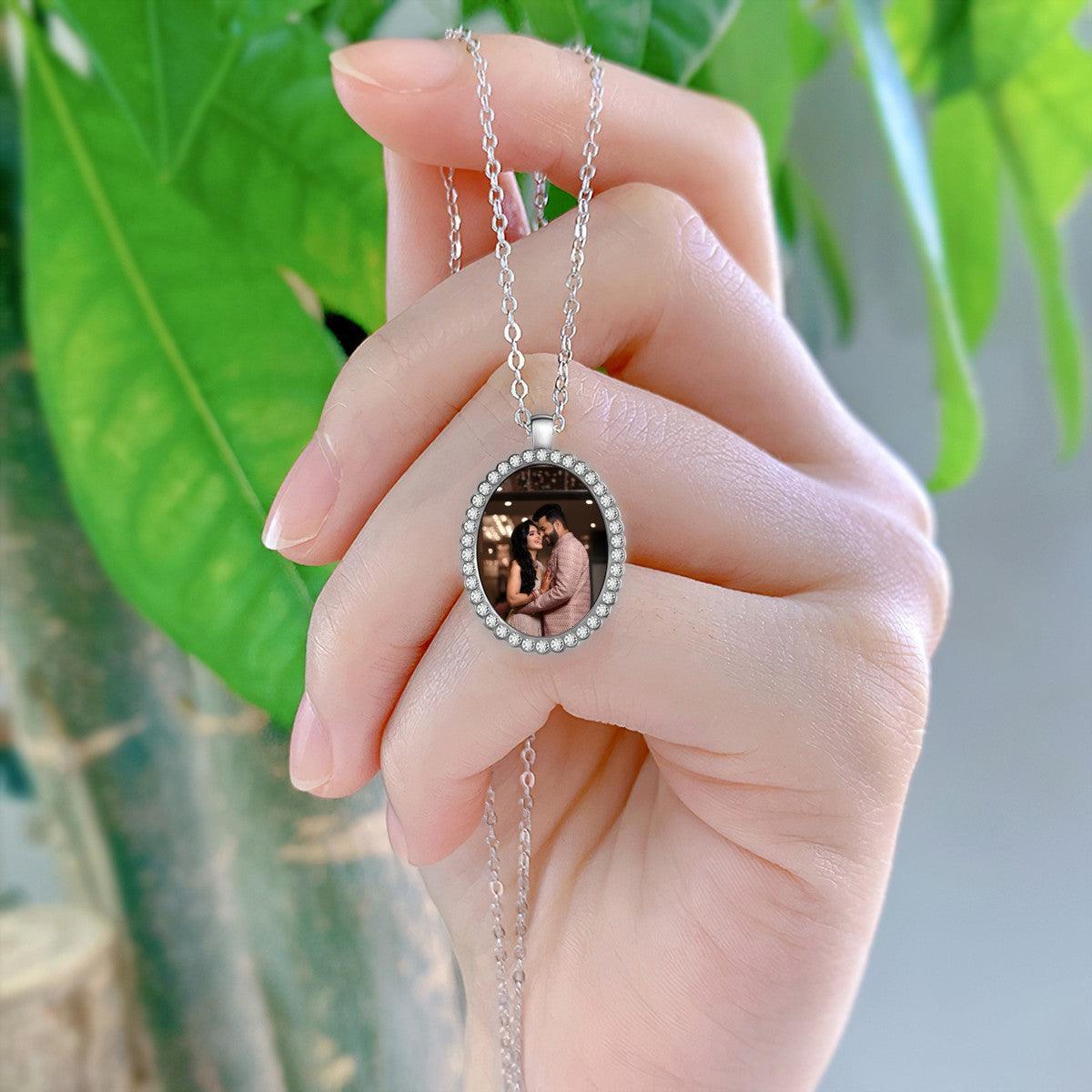 Silver oval photo pendant necklace held in a hand, with crystal border and couple photo