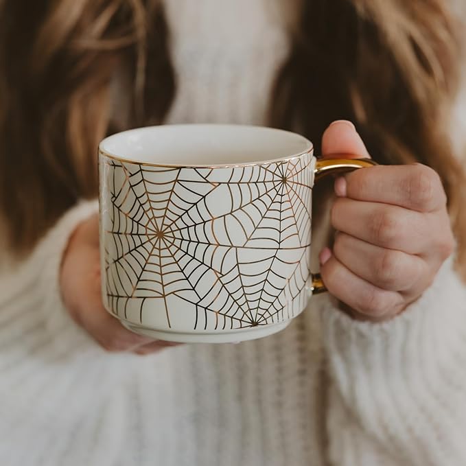 White mug with black spiderweb print and gold handle, held near chest