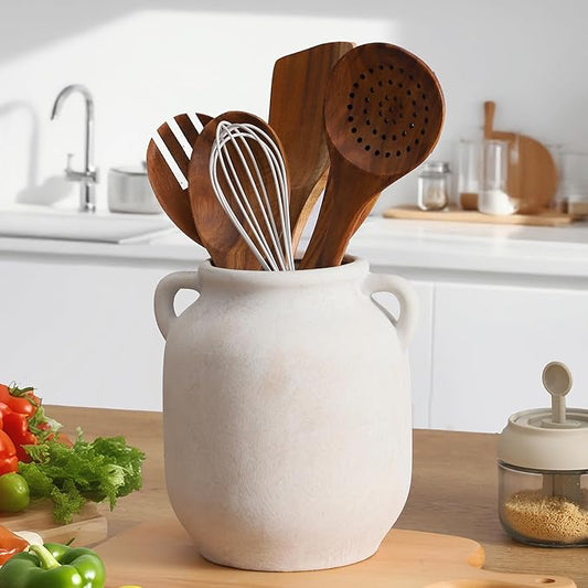 Rustic white ceramic vase with side handles holding kitchen utensils and whisk.