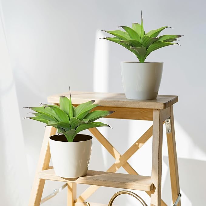 Two dark green faux agave plants in white pots on a wooden stand