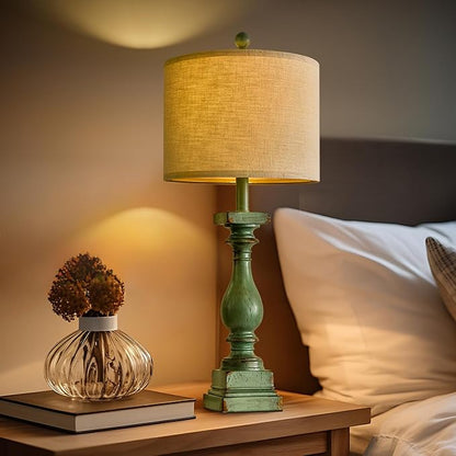 Green resin table lamp with beige drum shade on nightstand beside pillow