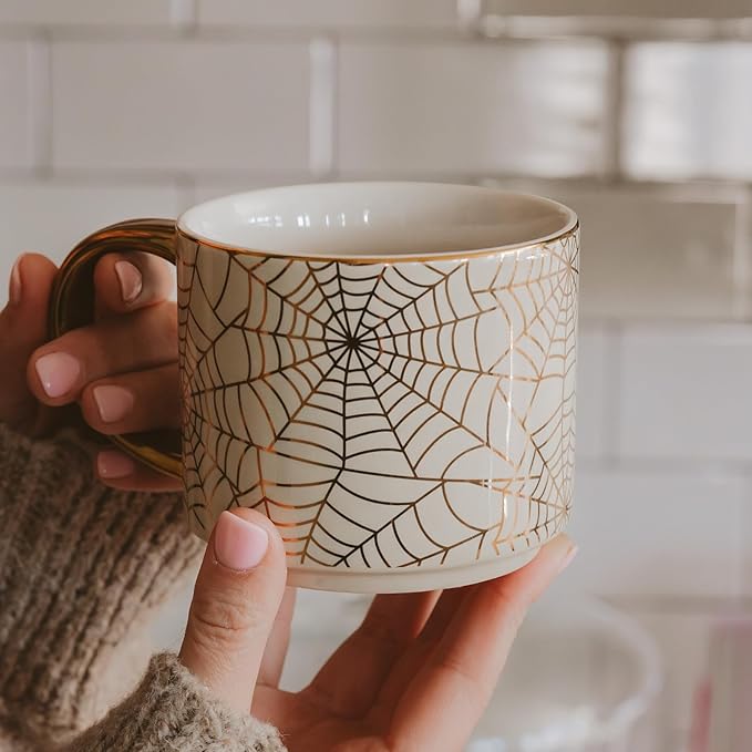 White mug with black spiderweb pattern and gold handle, held in a hand