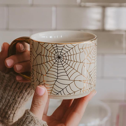 White mug with black spiderweb pattern and gold handle, held in a hand