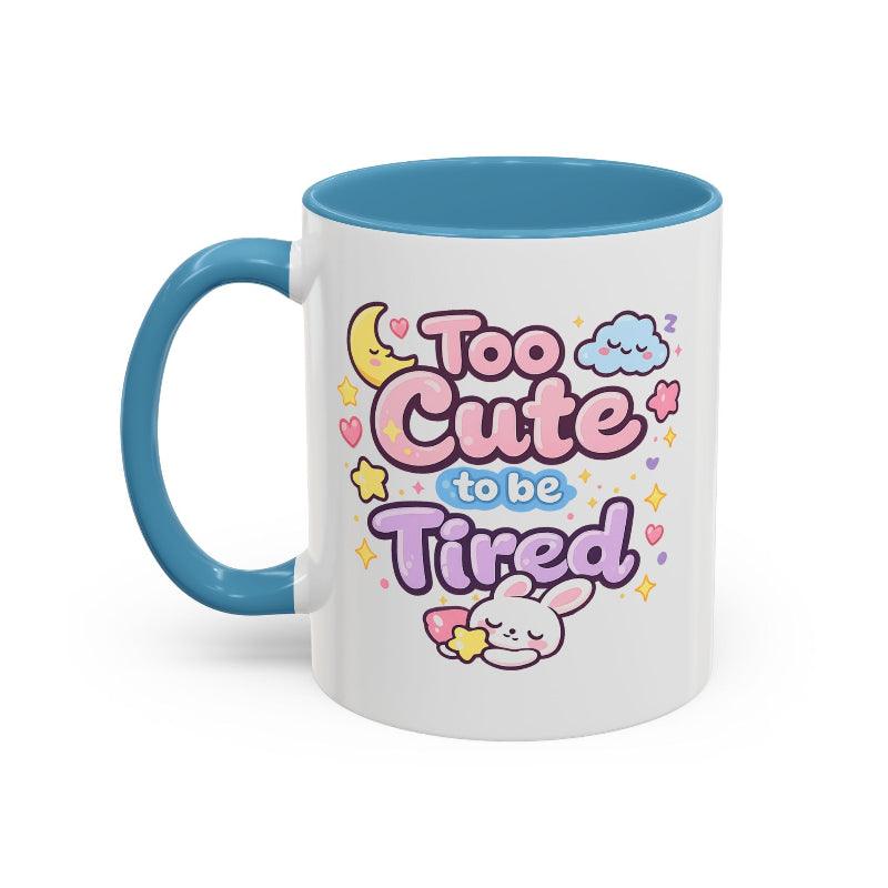 White mug with blue rim and handle, “Too Cute to be Tired” bunny graphic.