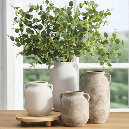 White ceramic vase with side handles holding leafy green stems on wooden stand