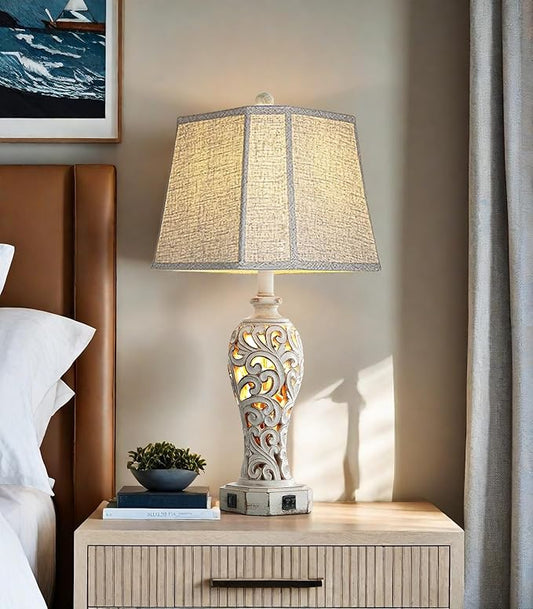 Farmhouse table lamp with beige fabric shade and ornate white resin base on end table.