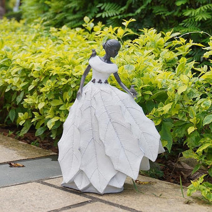 White resin fairy maiden statue beside green plants with layered skirt and bird figure