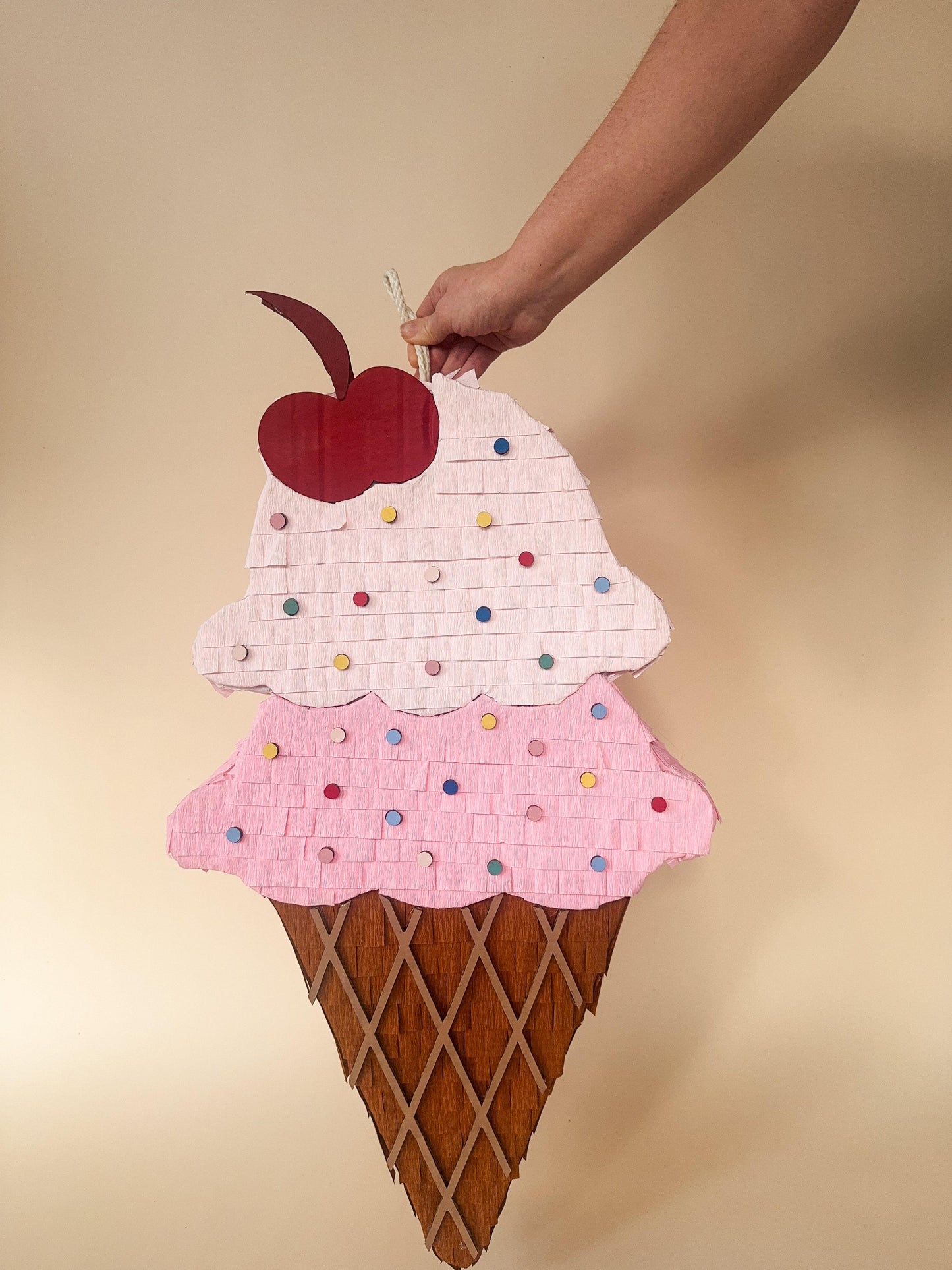 Pink ice cream sundae piñata held by hand, red cherry and colorful sprinkles on brown cone