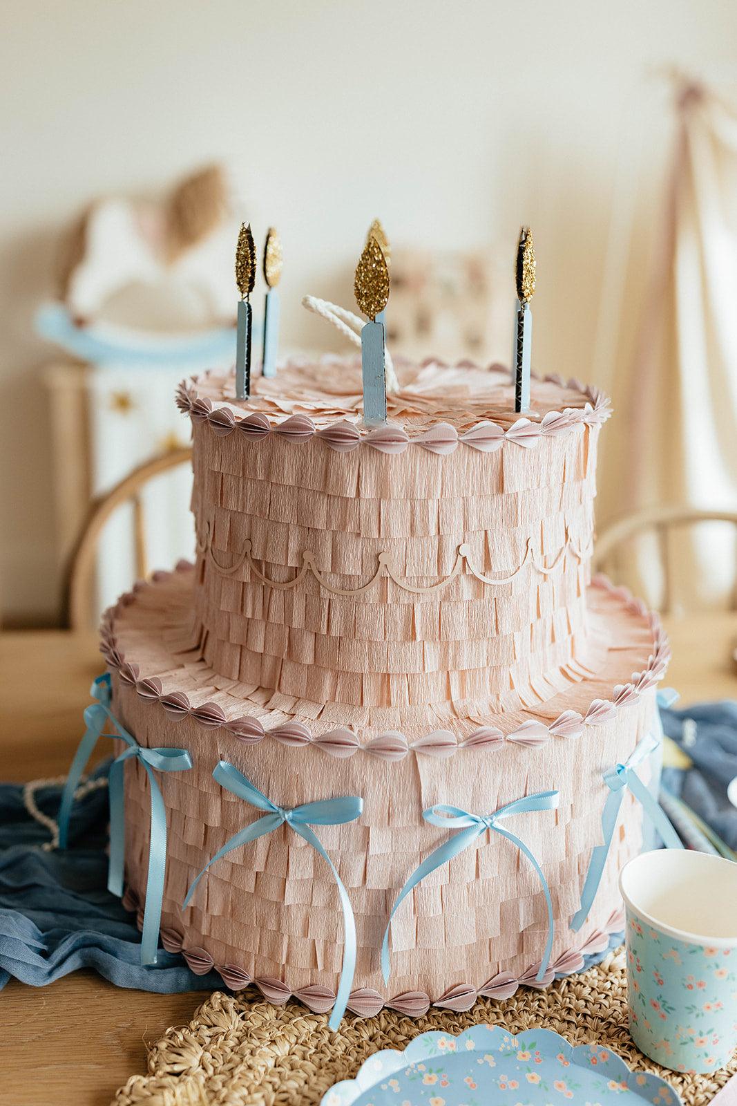 Piñata pastel rosa con velas negras y doradas, lazos azules y bordes con pliegues