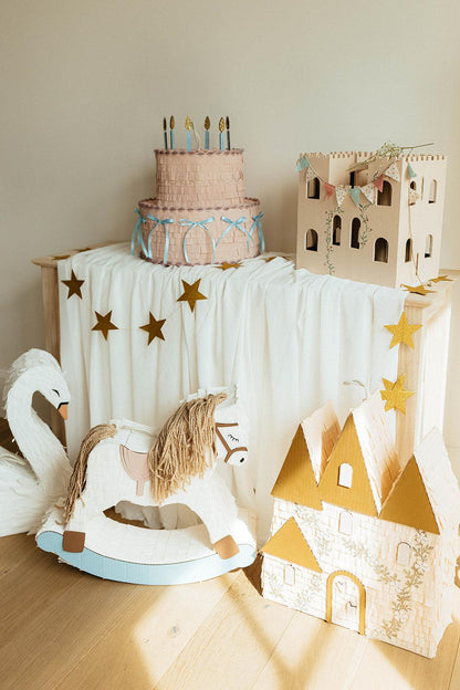Piñata pastel rosa con estrellas doradas, velas y cintas azules sobre mesa blanca