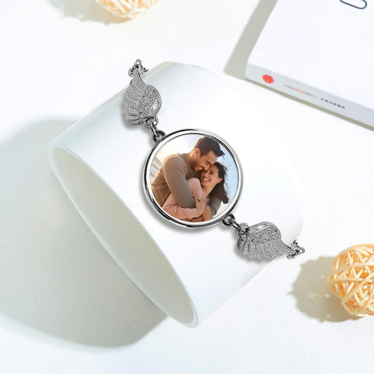 Silver photo charm bracelet with round locket and wing charms, laid on surface