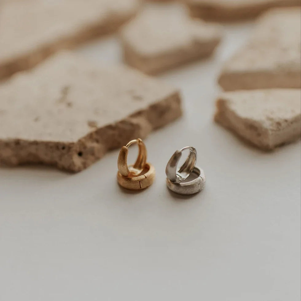 Chunky brushed huggie hoop earrings in gold and silver, shown open with hinged clasps.