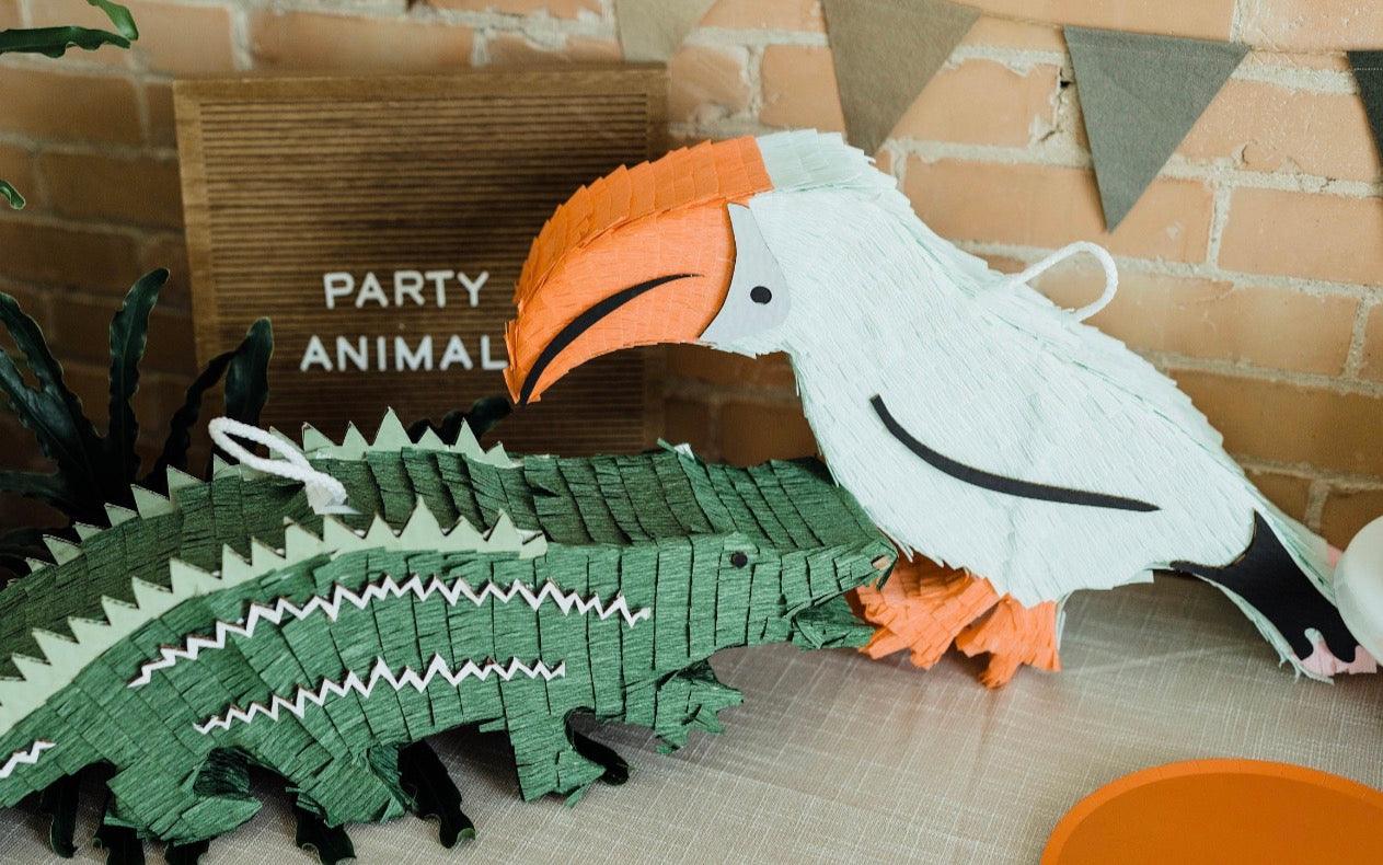 Tropical toucan piñata with white body, orange beak, and black smile, over green pieces