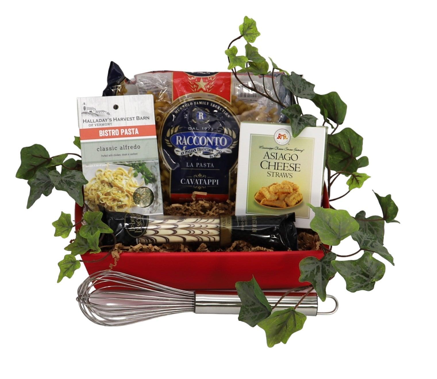 Red gift tray with pasta packages, cheese straws, and metal whisk, plus green leaves