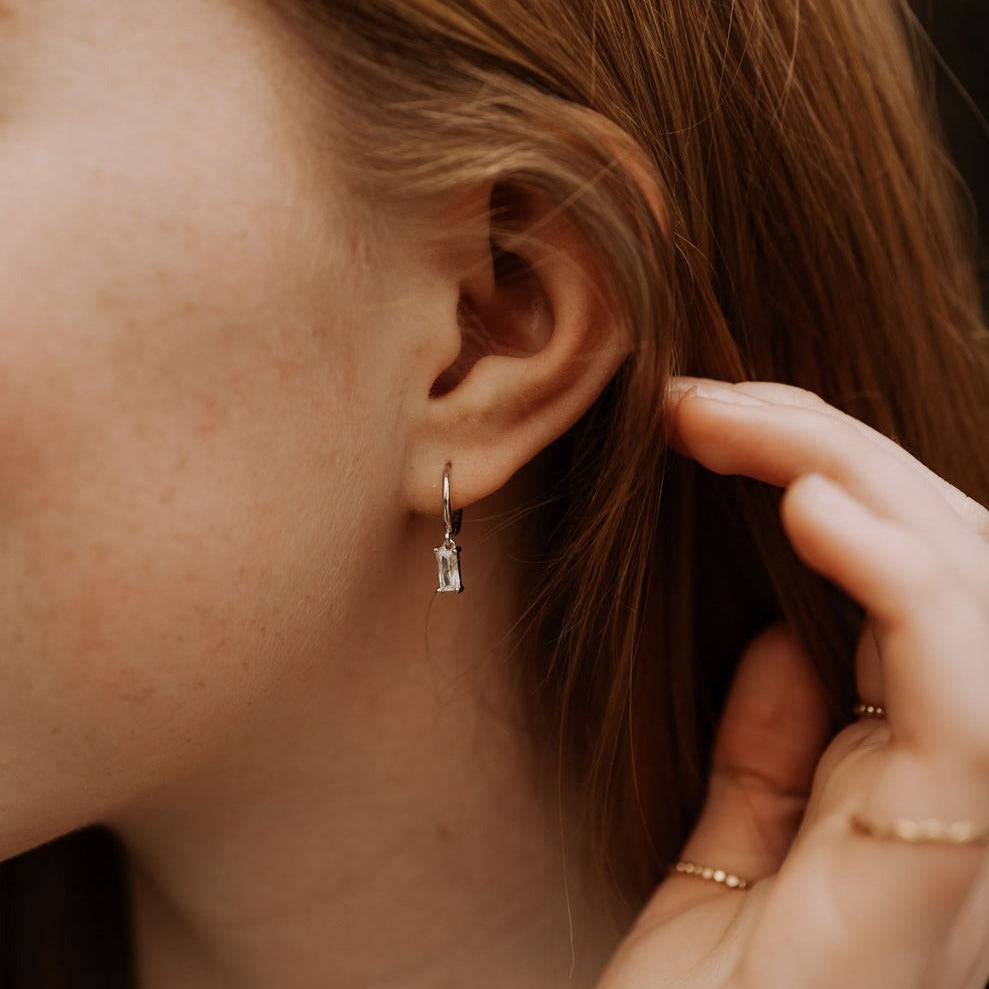 Silver hoop earring with a clear baguette-cut drop, worn on the ear