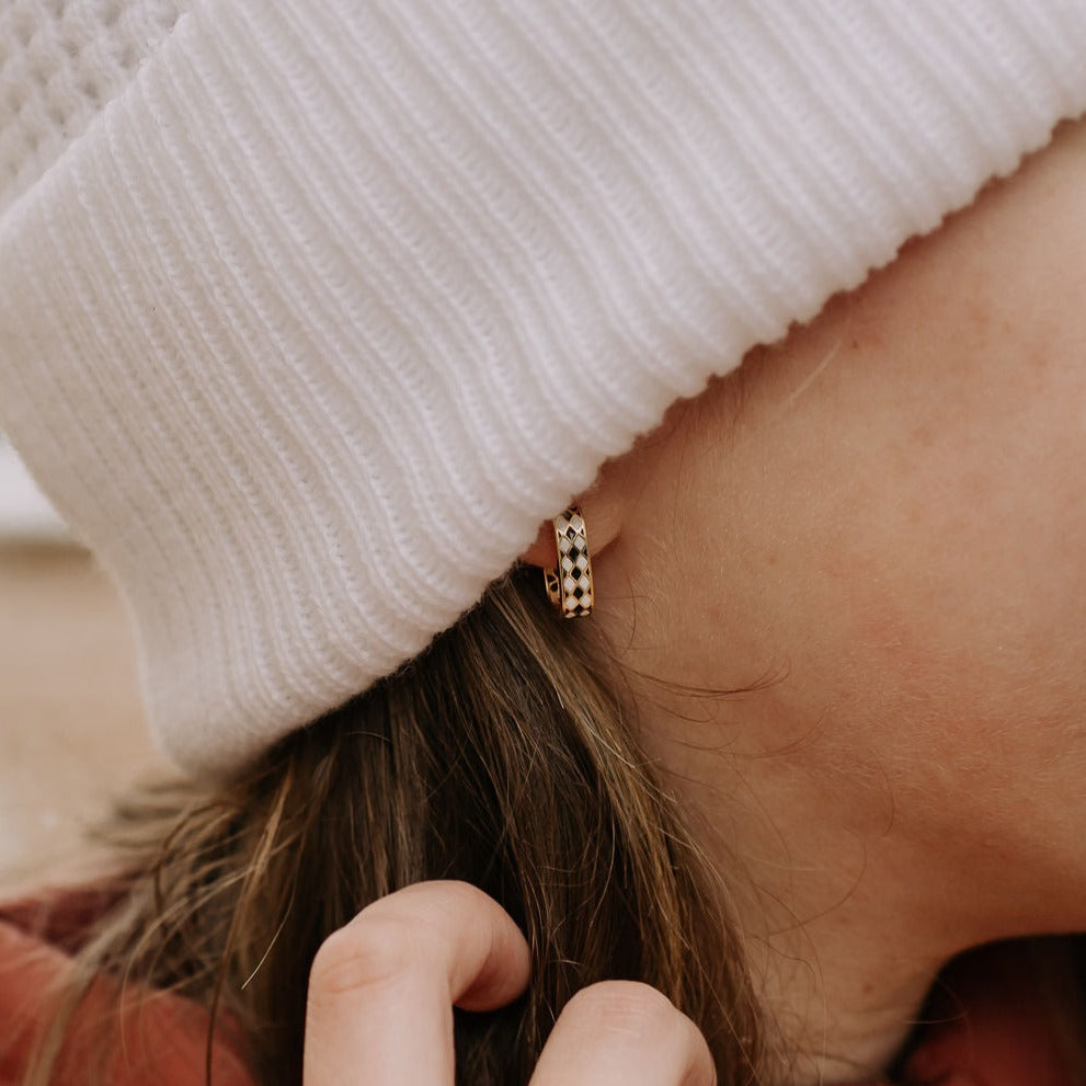 Black-and-white diamond-patterned gold hoop earring worn beneath a knit hat