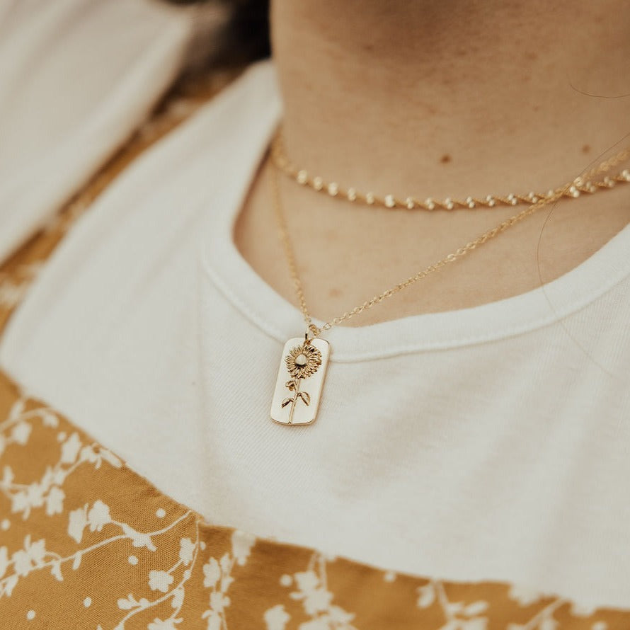 Gold necklace with a sunflower-engraved rectangular pendant, worn with a fine chain