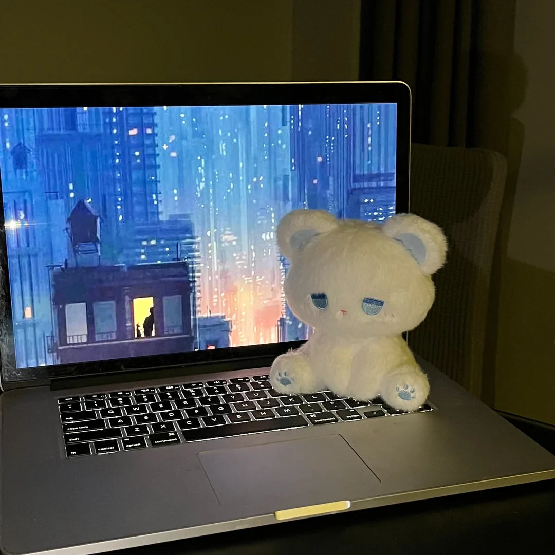 White plush cat with blue sleepy face sitting near a laptop, soft stuffed toy