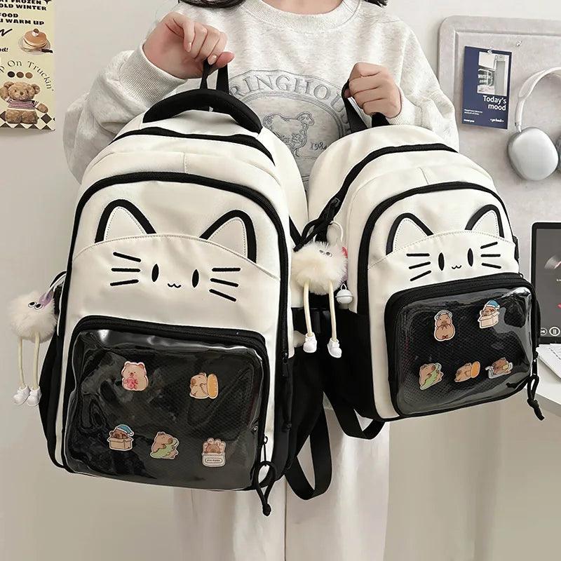 Two white cat backpacks with black trim; front clear pouch with colorful patches, straps on.