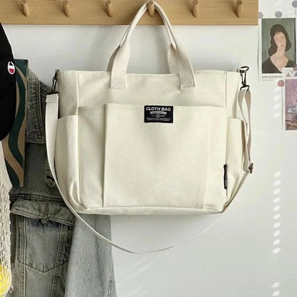 White canvas tote bag with two top handles and long crossbody strap, front zip pocket