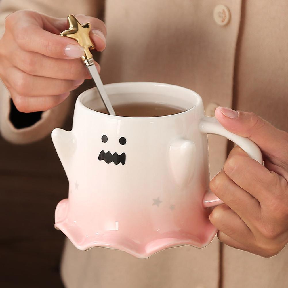 Ghost face mug with wavy mouth and gold star stirrer held near the handle; pastel pink base