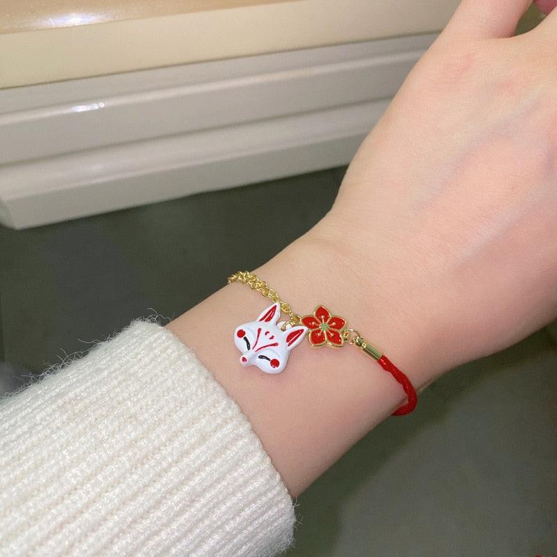 Red cord bracelet with gold chain, fox head charm, and red flower charm on wrist