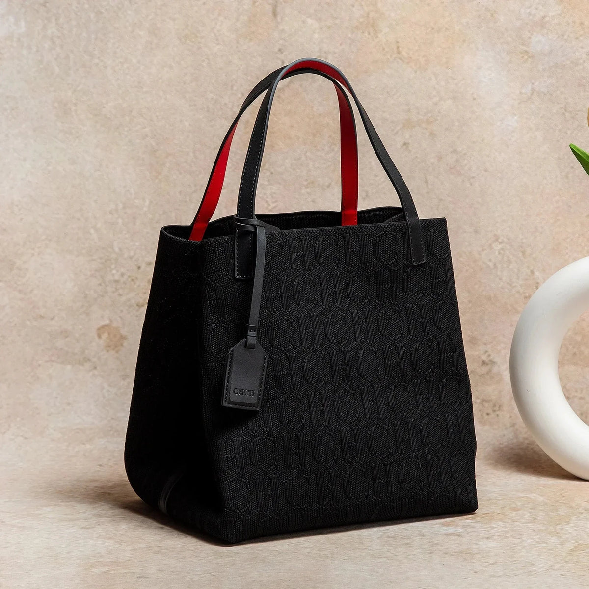 Black knitted tote bag with embossed CHCH pattern, red-lined handles, hanging tag.