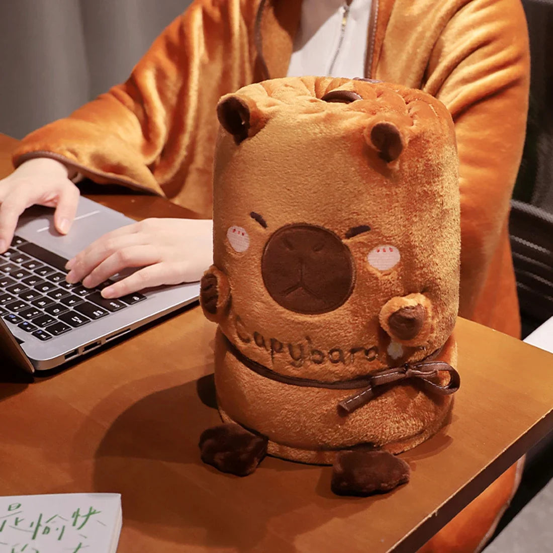 Rolled capybara-shaped blanket pillow, orange plush with stitched face and tie cord.
