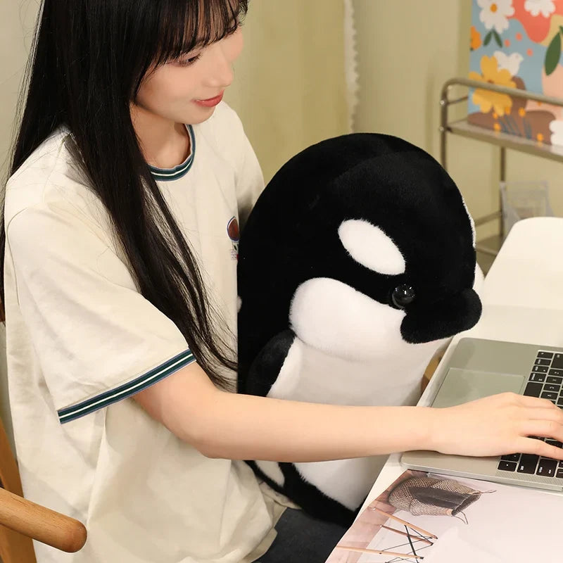 Black and white dolphin plush resting in arms next to a laptop on a desk.