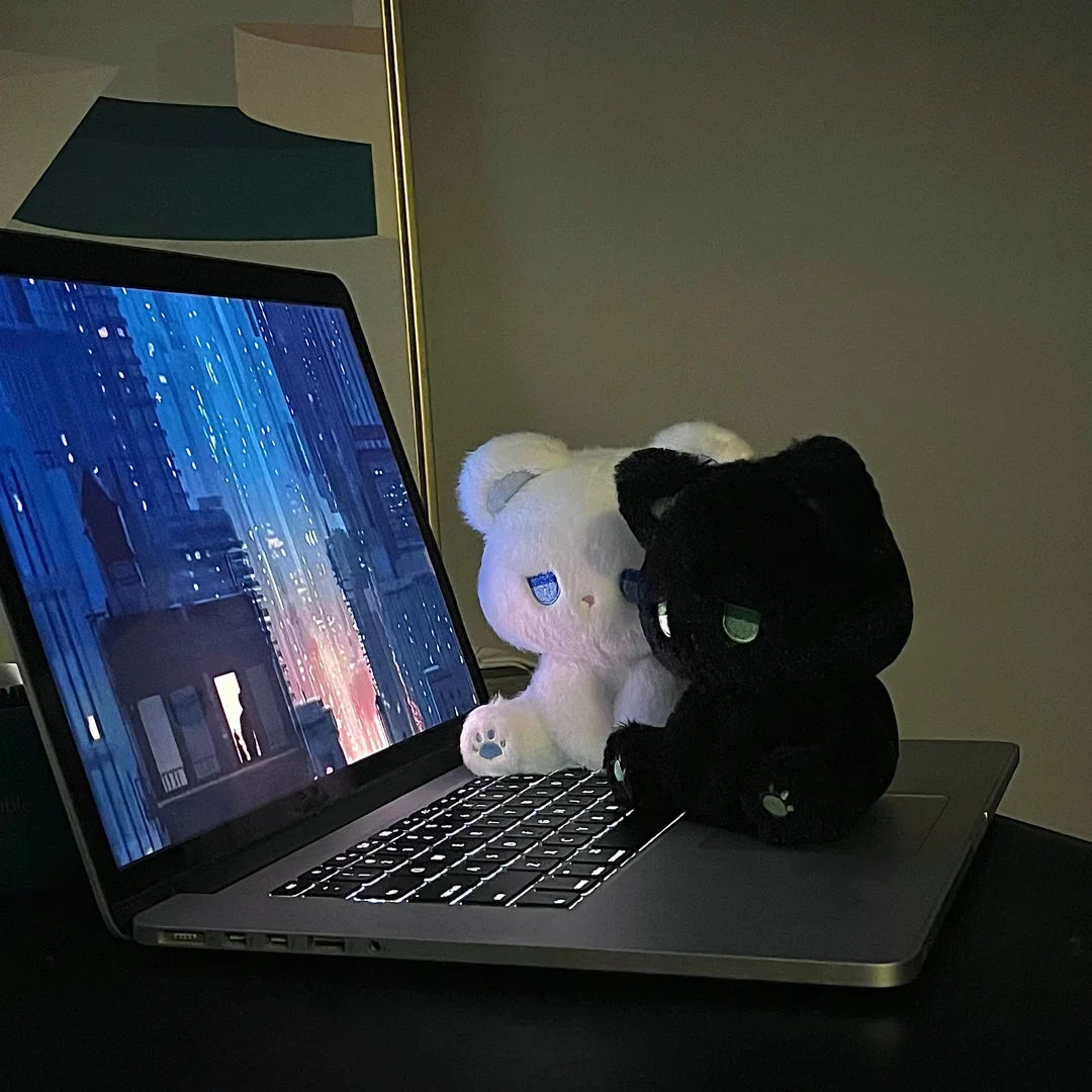White and black plush cats with sleepy faces sitting beside a laptop