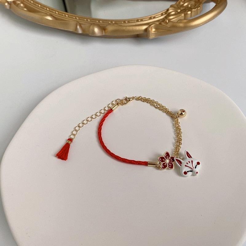 Fox charm bracelet with red cord, gold link chain, and bell detail laid on a white pad