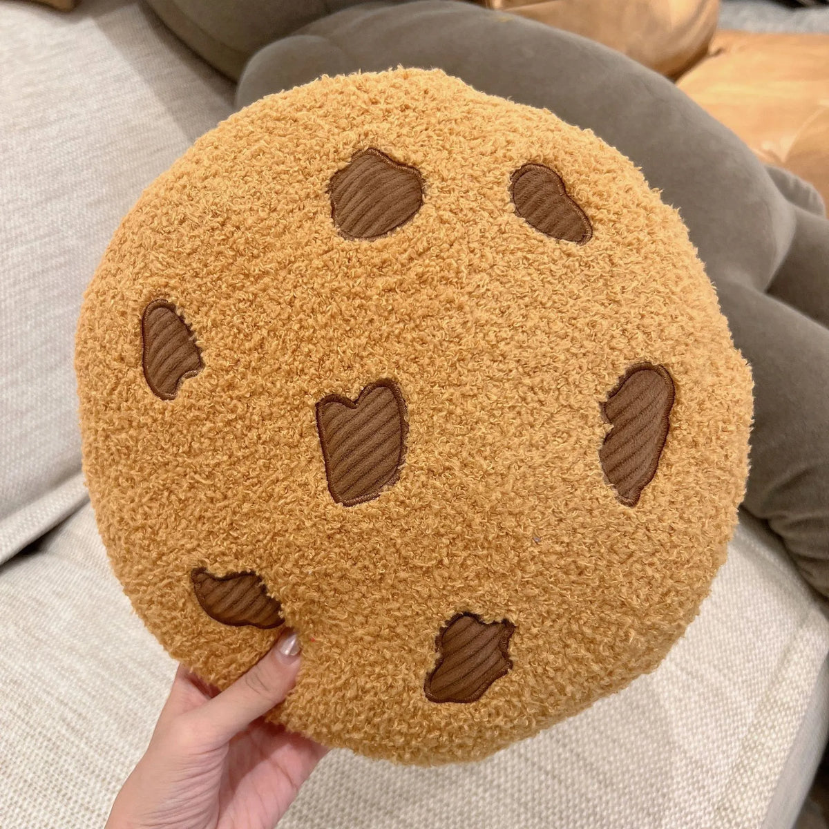 Round tan cookie plush pillow with brown stitched chocolate-chip shapes, held in hand