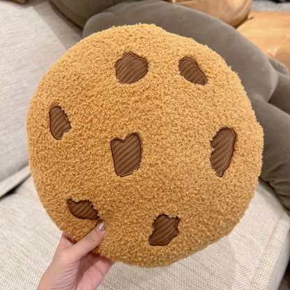 Round tan cookie plush pillow with brown stitched chocolate-chip shapes, held in hand
