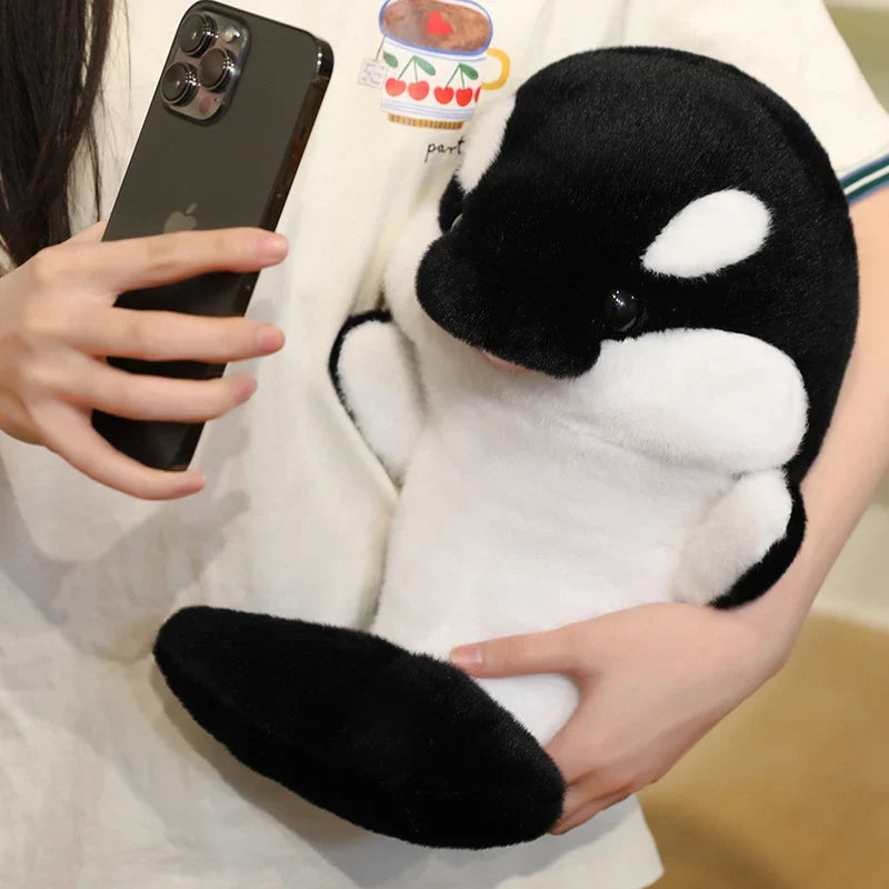 Black and white dolphin plush held up beside a phone, large oval white eye patch.
