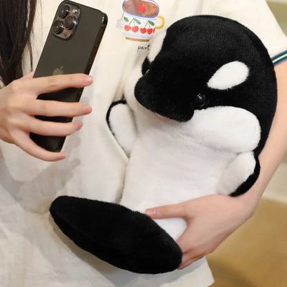 Black and white dolphin plush held up beside a phone, large oval white eye patch.