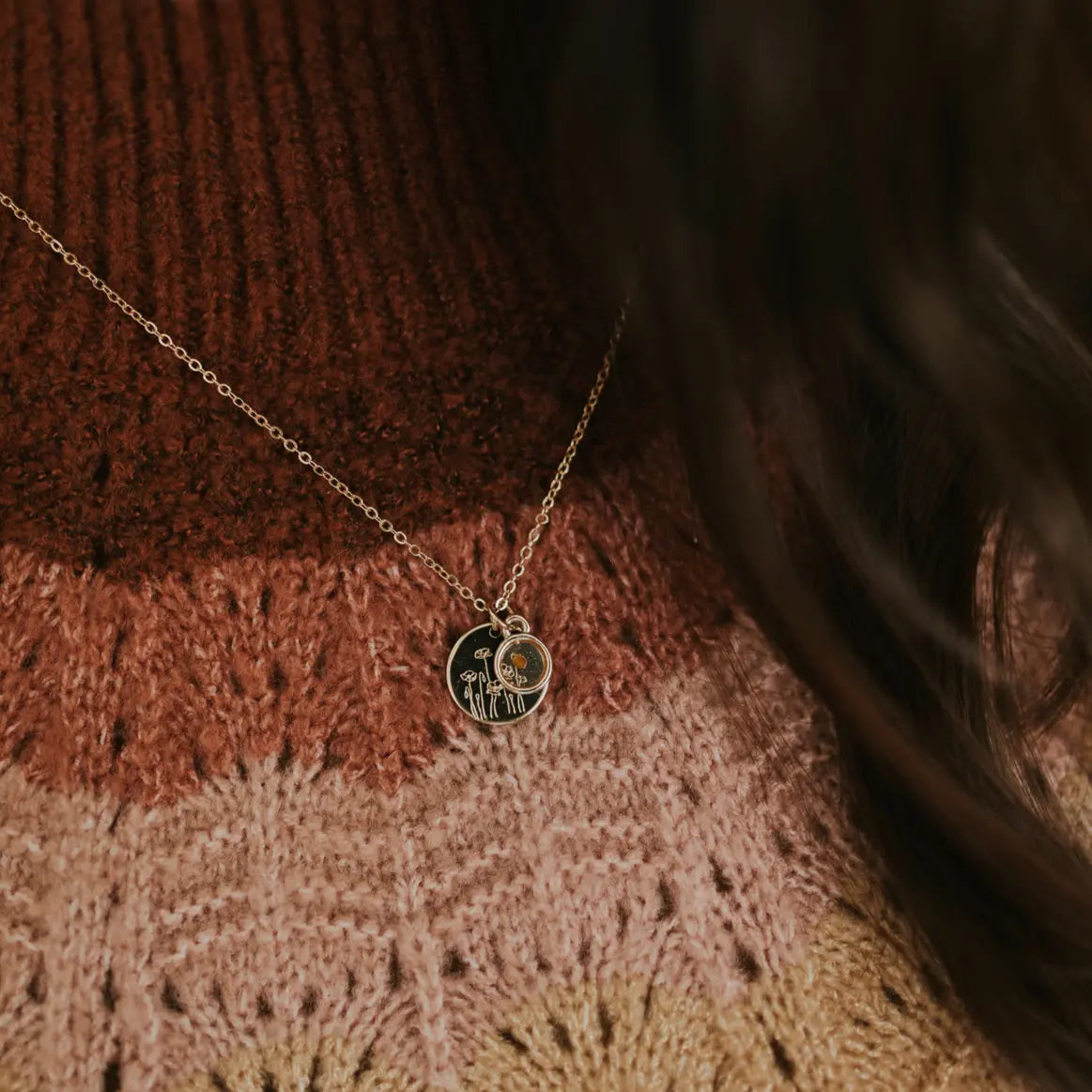 Gold-tone poppy necklace with engraved disc and small mustard seed charm, worn on a sweater