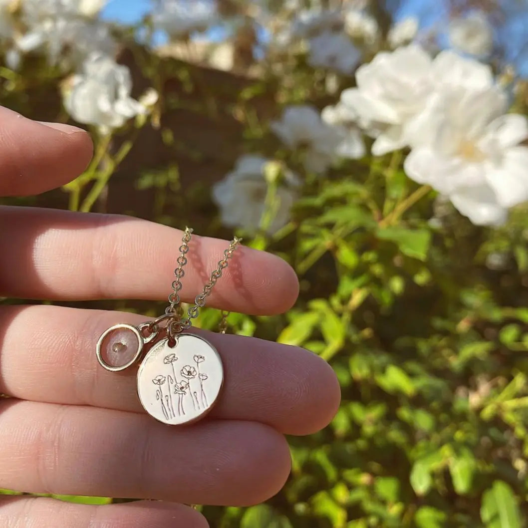 Gold-tone poppy necklace with engraved pendant and seed charm, held in a hand