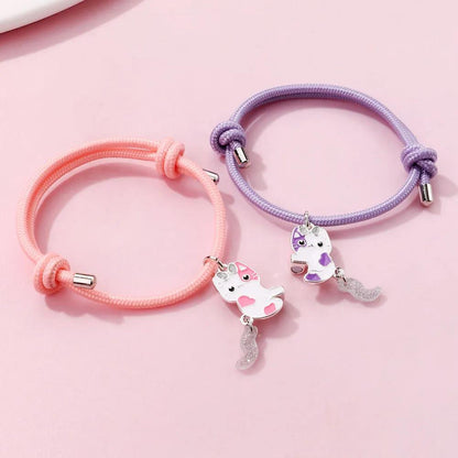 Two braided bracelets side by side: pink and lavender bands with cat charms and sparkly tassels