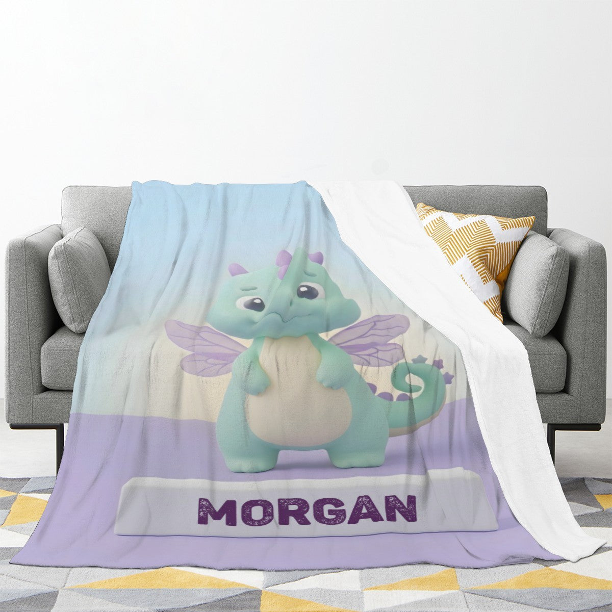 Printed blanket laid on sofa; light blue dragon design with purple bottom band and “MORGAN”